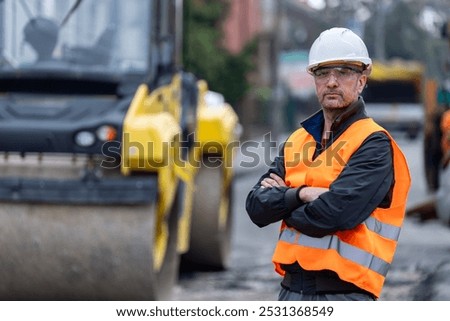 A construction worker wearing a hard hat and high-visibility vest stands confidently with crossed arms, monitoring heavy machinery at a bustling urban construction site in daylight.