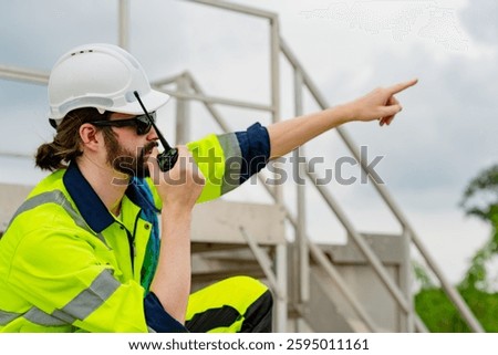 A construction worker wearing a hard hat and reflective suit uses a radio to coordinate tasks on-site. He points to direct attention, highlighting active teamwork during cloudy weather.