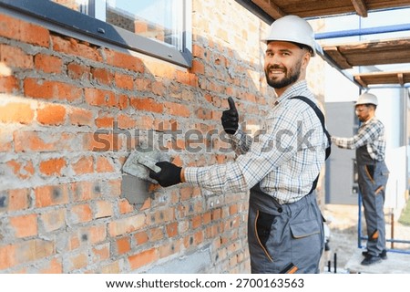 Construction worker wearing hard hat and uniform, smiling at camera, giving thumbs up while plastering brick wall