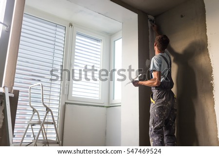 Construction worker wearing worker overall with wall plastering tools renovating apartment house. Plasterer renovating indoor walls and ceilings with float and plaster. Construction finishing works.