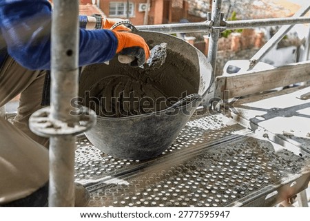 A construction worker, wearing protective gloves, mixing cement in a bucket on scaffolding