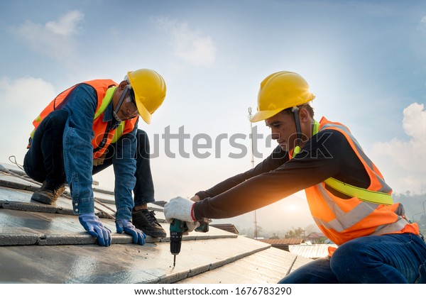 Construction worker wearing safety harness belt\
during working on roof structure of building on construction\
site,Roofer using air or pneumatic nail gun and installing concrete\
roof tile on top\
roof.