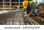 Construction worker wearing a yellow hard hat and safety vest, kneeling and working on steel rebar at a construction site.