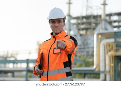 A construction worker in a white hard hat and orange safety jacket points directly at the camera at an industrial site.
