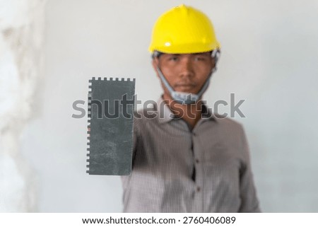 A construction worker in a yellow hard hat is holding a trowel.