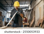 Construction worker in a yellow hardhat and safety glasses inspecting a board of wood. Ideal for carpentry, woodworking, and quality control, highlighting craftsmanship in the workshop. Labor day