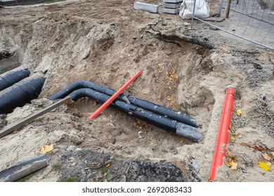 Construction workers are digging a trench to install large underground pipes made of plastic and metal in a residential area. Leaves are scattered on the ground.