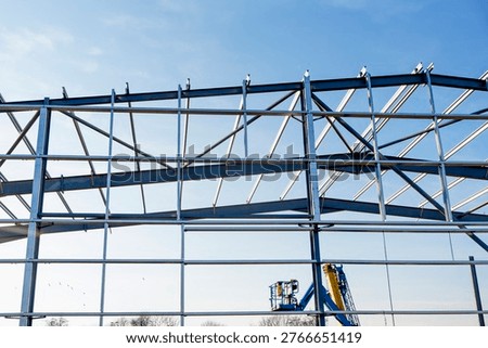 Construction workers are erecting a steel framework at a building site during the day. Machinery is visible in the background, and blue sky overhead completes the scene.