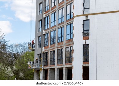 Construction workers installing facade insulation and finishing exterior of modern apartment building. Residential construction site with unfinished architecture and suspended platforms.