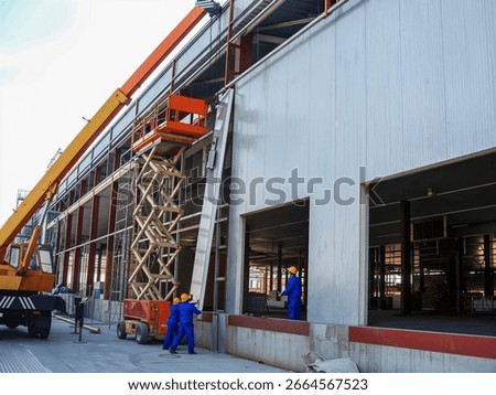 Construction workers installing facade panels on a building using a lift, with a crane nearby, capturing an active construction site.