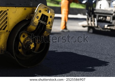 Construction workers operate machinery to compact freshly laid asphalt on a roadway during the afternoon. The process ensures surface durability for traffic.
