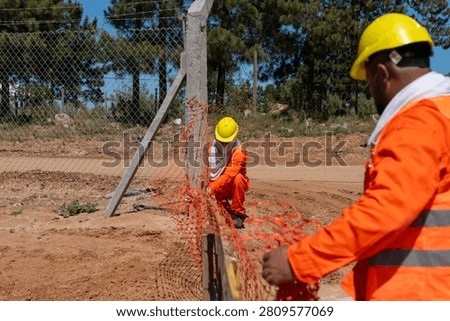 Construction Workers in Orange Uniforms and Hard Hats Installing Safety Barrier Netting at Work Site