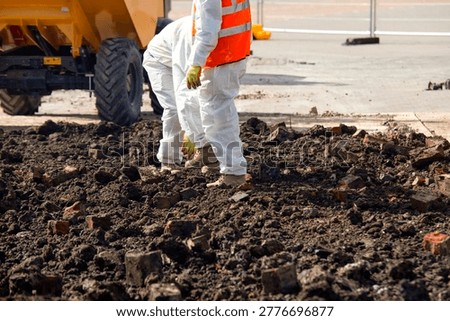 Construction workers are seen on a construction site during the afternoon. They are moving through dirt and bricks while machinery operates in the background.