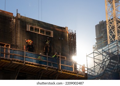 Construction Workers Seen At Work On The Side Of A Under Construction Building Frame As The Sun Shines Through A Clear Day.