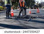 Construction workers using a jackhammer to dig up freshly laid asphalt to find the sewar access point below and raise the entrance up to the new street level, road repaving project
