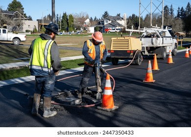 Construction workers using a jackhammer to dig up freshly laid asphalt to find the sewar access point below and raise the entrance up to the new street level, road repaving project
