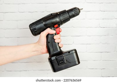 Construction Working Tool, Electric Screwdriver In A Female Hand On The Background Of A White Brick Wall. Cordless Drill With A Battery Pack In Hand Close-up