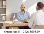 Consul at Consulate of United States of America sitting at office table with American flag and listening to young man telling about his problems with immigration and USA travel visa application