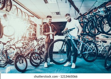 Consultant Shows Bicycle to Client in Sport Shop. Portrait of Young Shop Assistant Wearing White T-Shirt Helps in Mountain Bike Choosing. Happy Salesman in Sport Store with Row of Bikes on Backround - Powered by Shutterstock