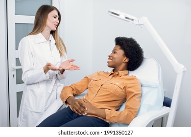 Consultation in cosmetology clinic. Female professional beauty doctor talking with pretty smiling African woman patient and telling good news about skin care and treatment. Skin care and cosmetology.