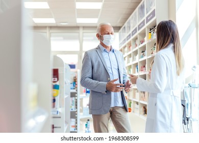 Consultation With A Pharmacist. A Mature Gray-haired Man Wears A Protective Mask And Stands In The Middle Of The Pharmacy And Talks To A Female Pharmacist In Uniform. Pleasant Conversation And Smiling