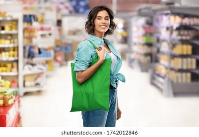 Consumerism, Sustainability And People Concept - Portrait Of Happy Smiling Young Woman In Turquoise Shirt With Green Reusable Canvas Bag For Food Shopping Over Supermarket Or Grocery Store Background