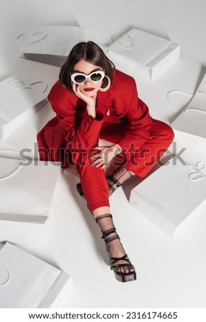 consumerism, young woman with brunette short hair, nose piercing and tattoo posing in sunglasses and red suit while sitting around shopping bags on grey background, high angle view