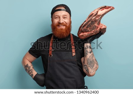 Consumption concept. Cheerful friendly ginger meatpacking worker has manufacturing job, holds slaughtered raw meat, transforms ingredients into food at factory, wears rubber gloves and apron