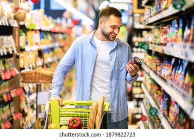 Consumption Concept. Portrait Of Smiling Bearded Guy With Shopping Cart In Market Buying Groceries Food Walking Along The Aisle And Shelves In Store, Holding Glass Jar Of Sauce, Checking Label