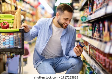 Consumption And Consumerism. Portrait Of Smiling Bearded Man With Shopping Cart In Market Buying Groceries Food Taking Products From Shelves In Store, Holding Glass Jar Of Sauce, Checking Label