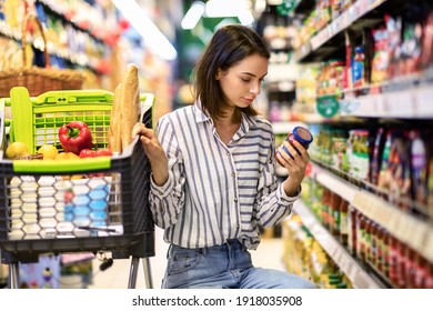 Consumption And Consumerism. Portrait Of Young Woman With Shopping Cart In Market Buying Groceries Food Taking Products From Shelves In Store, Holding Glass Jar Of Sauce, Checking Label Or Expiry Date