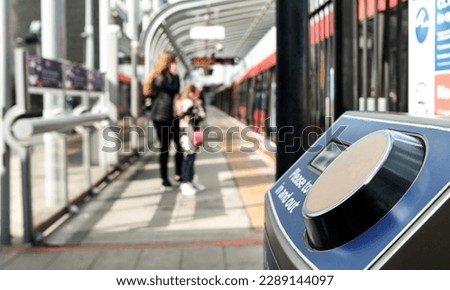 Contactless card reader machine for public transport in London. There are a red train and people out of focus in the background. Public transport and technology concept.