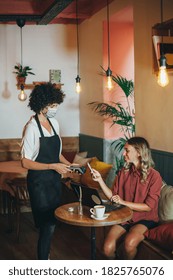 Contactless Payment With Mobile In Coffee Shop. Waitress Standing, Wearing Protective Face Mask And Customer Sitting With Her Mobile On Hand About To Pay With Just A Tap During Coronavirus Pandemic