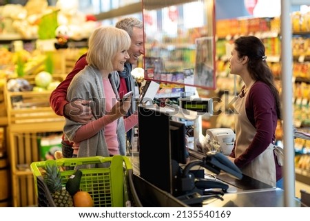 Contactless payment at supermarket concept. Happy loving elderly husband and wife paying at cashdesk, cheerful senior man and woman having conversation with cashier while paying with smartphone