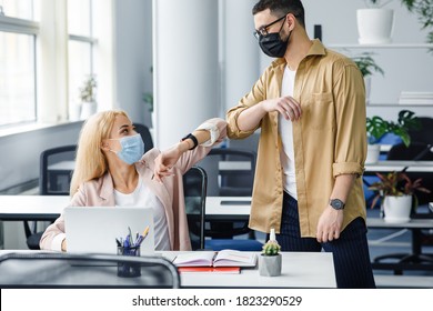 Contactless To Say Hello, Health Care And Social Distance During Covid-19 Pandemic. Young Man And Lady In Protective Masks Greet With Elbows At Workplace In Office Interior, Empty Space