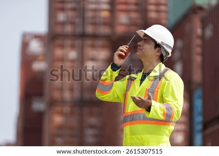 container loading worker in fluorescent safety jacket and hard hat is using walkie-talkie radio to communicate while surrounded stacked shipping containers. His role involves coordinating logistics.