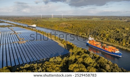 Container ship and car ferry navigate the Kiel Canal alongside vast solar panel fields and wind turbines, set against a backdrop of forest.