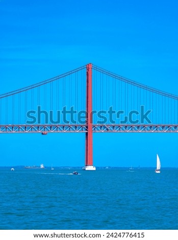 Container ship, motorboats and sailboats navigating the Tagus River crossing the red steel 25 de Abril suspension bridge with road traffic for access to Lisbon, under a clear blue sky.