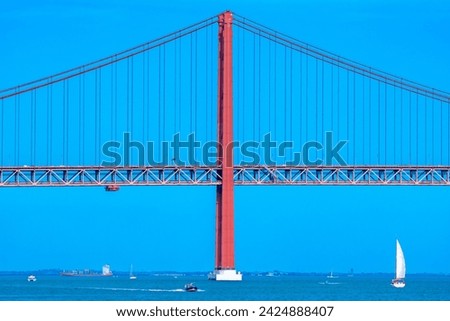 Container ship, motorboats and sailboats navigating the Tagus River crossing the red steel 25 de Abril suspension bridge with road traffic for access to Lisbon, under a clear blue sky.