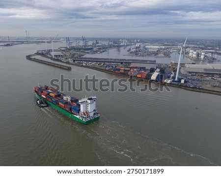Container ship on river Thames escorted by pilot boat. Tilbury docks with wind turbines. High altitude aerial view on a cloudy day. Cargo and shipping on the outskirts of London, UK.