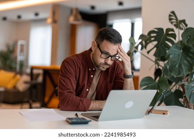 Contemplating businessman in the home office feeling frustrated. Full concentration on work. Man working on laptop, counting profit while sitting at the desk at home. - Powered by Shutterstock