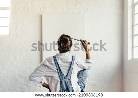 Contemplating creative ideas for a new project. Rearview of a young painter looking thoughtful while sitting in front of a blank canvas. Creative female artist holding a paintbrush to her head.
