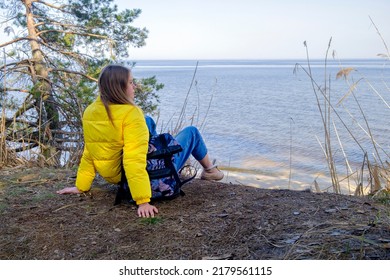 Contemplation. Young Woman With Backpack Is Sitting On The Coast Of Forest Lake At Cloudy Day In Spring. Time For Yourself. Contemplation And Rest In Solitude.Landscape With Slim Girl, Green Trees.