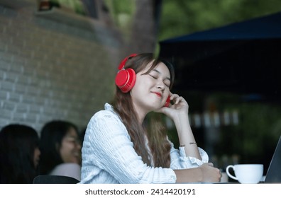 A contemplative Asian woman listens to music on red headphones while working on her laptop in a cozy café setting.