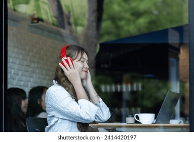A contemplative Asian woman listens to music on red headphones while working on her laptop in a cozy café setting.
