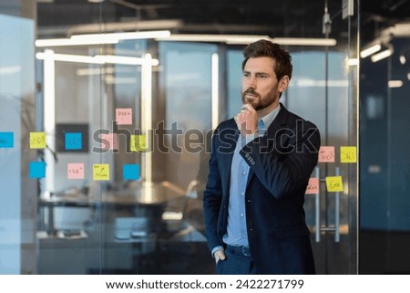 A contemplative businessman in a suit stands pondering before a glass wall filled with sticky notes and data charts.