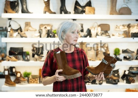 Contemplative elderly woman browsing through diverse models of leather and suede womens boots in shoe store while choosing demi-season footwear..
