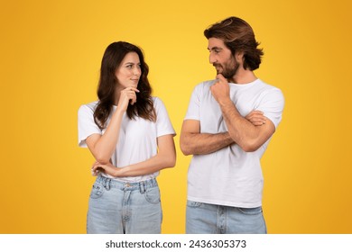 A contemplative european young couple in white t-shirts and jeans, looking at each other with thoughtful expressions, suggesting deep conversation or decision-making, against a yellow background - Powered by Shutterstock