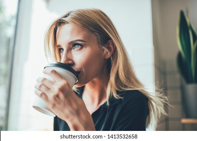 Contemplative hipster girl with blonde hair thoughtful looking away while drinking tasty caffeine beverage from takeaway cup, attractive pensive woman holding coffee and enjoying good taste