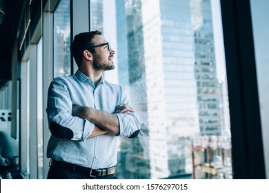 Contemplative Male Entrepreneur With Crossed Hands Standing Near Office Window View And Feeling Pondering During Work Day In Company, Caucasian Pensive Corporate Boss Thoughtful Looking Away